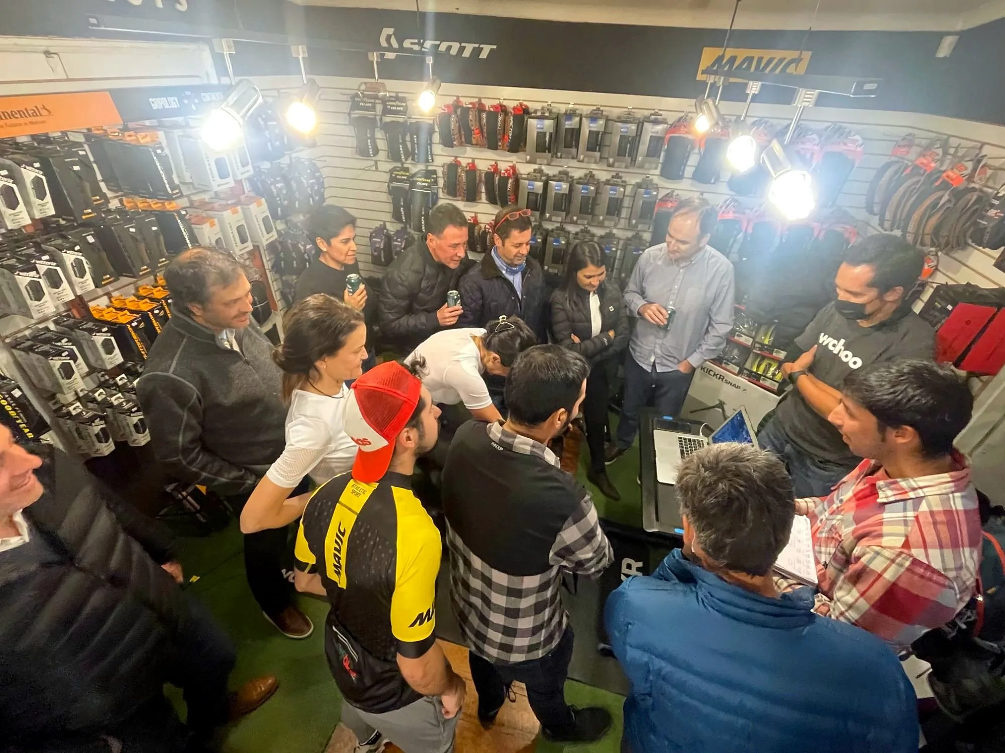 Grupo de ciclistas reunidos en una tienda de bicicletas, entre muros de neumáticos, mirando una presentación en un notebook
