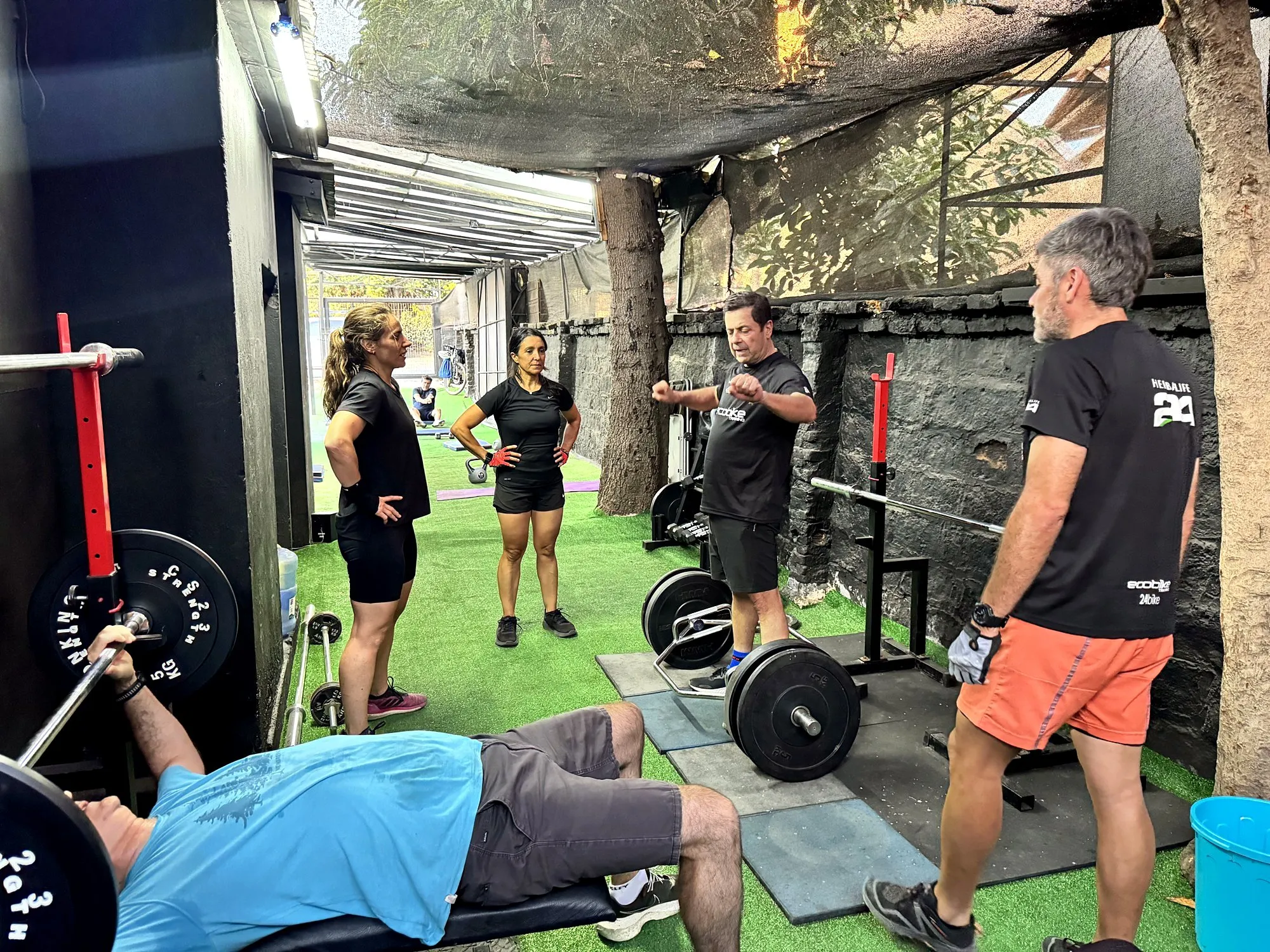 Grupo de ciclistas haciendo trabajo de fuerza en un gimnasio, con barras y discos, mientras un entrenador da indicaciones