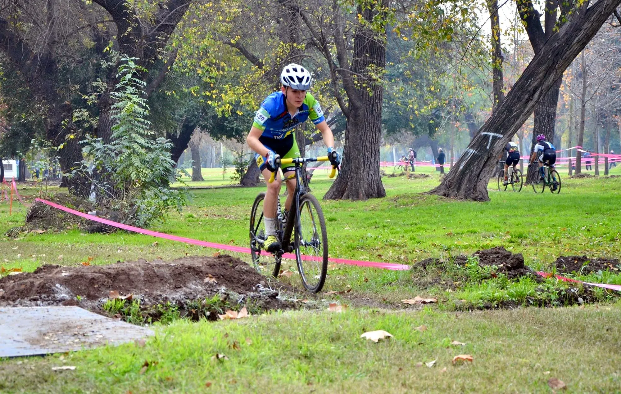 Corredor juvenil con dorsal en una carrera de ciclocross en un parque, pasando junto a un montículo de tierra entre árboles, con otros corredores al fondo