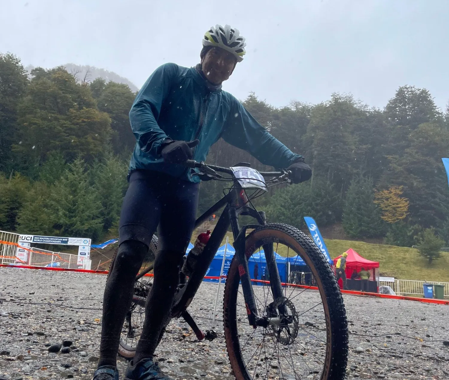 Marcelo, de Mundobikes, con chaqueta impermeable y guantes junto a su MTB embarrada, bajo la lluvia en el Mundial UCI Masters de MTB 2022 en Cerro Bayo, Argentina