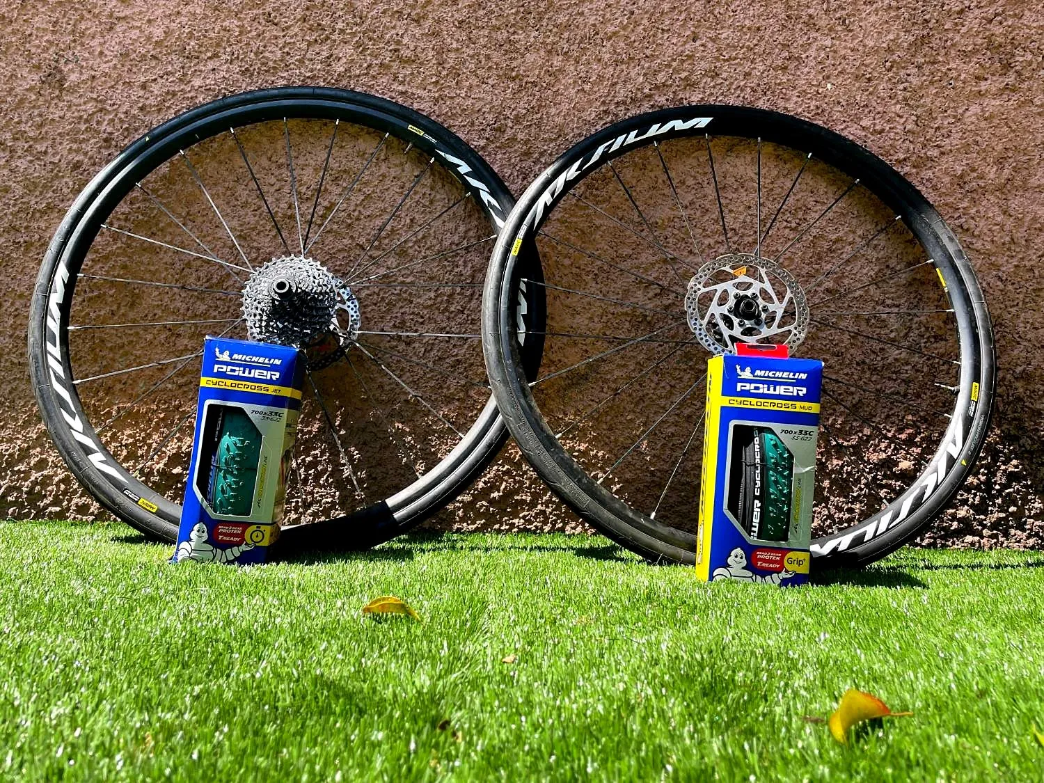Dos ruedas de ciclocross apoyadas en un muro con cajas de neumáticos Michelin Power Cyclocross Jet, para seco, y Mud, para barro, de 33 mm
