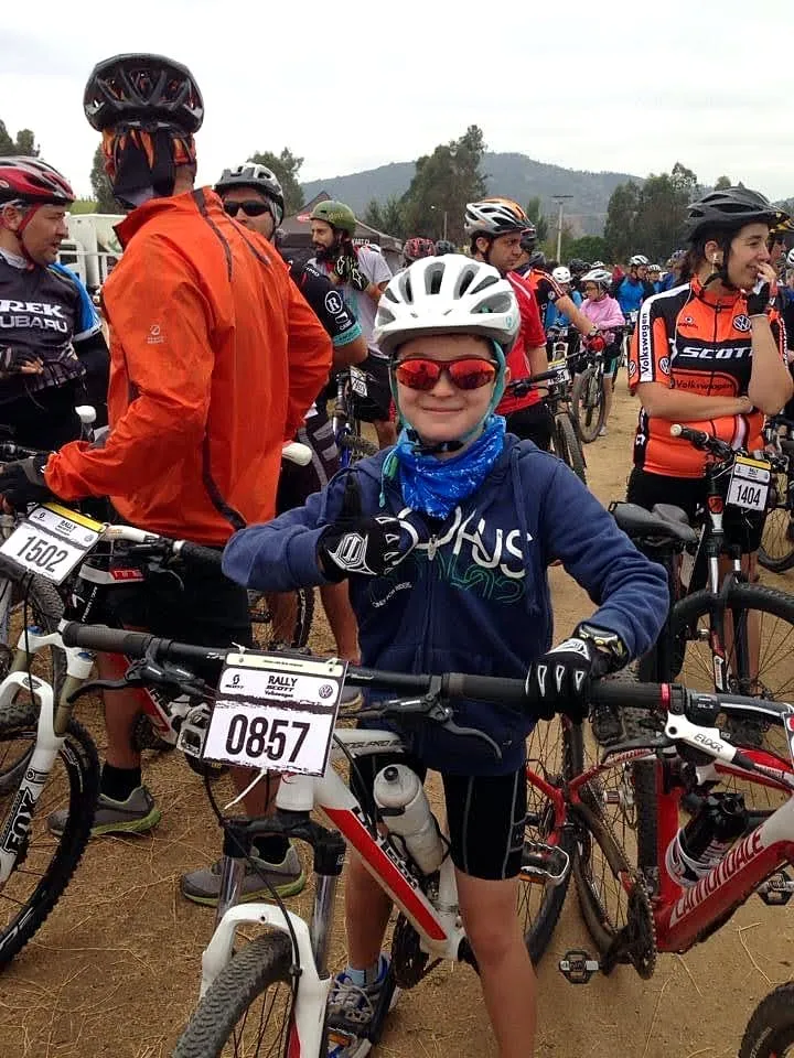 Niño con casco, lentes y dorsal en su bici de montaña, sonriendo en la zona de largada de una carrera de MTB rodeado de otros corredores