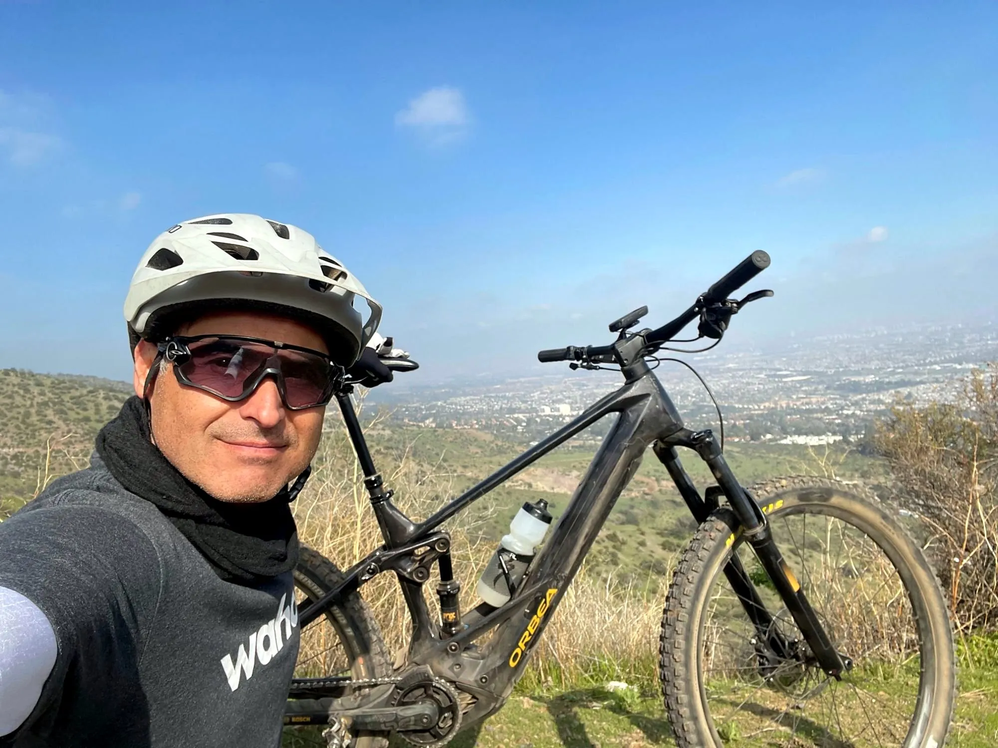Ciclista con casco y lentes junto a su eMTB Orbea Wild en un mirador, con Santiago de fondo