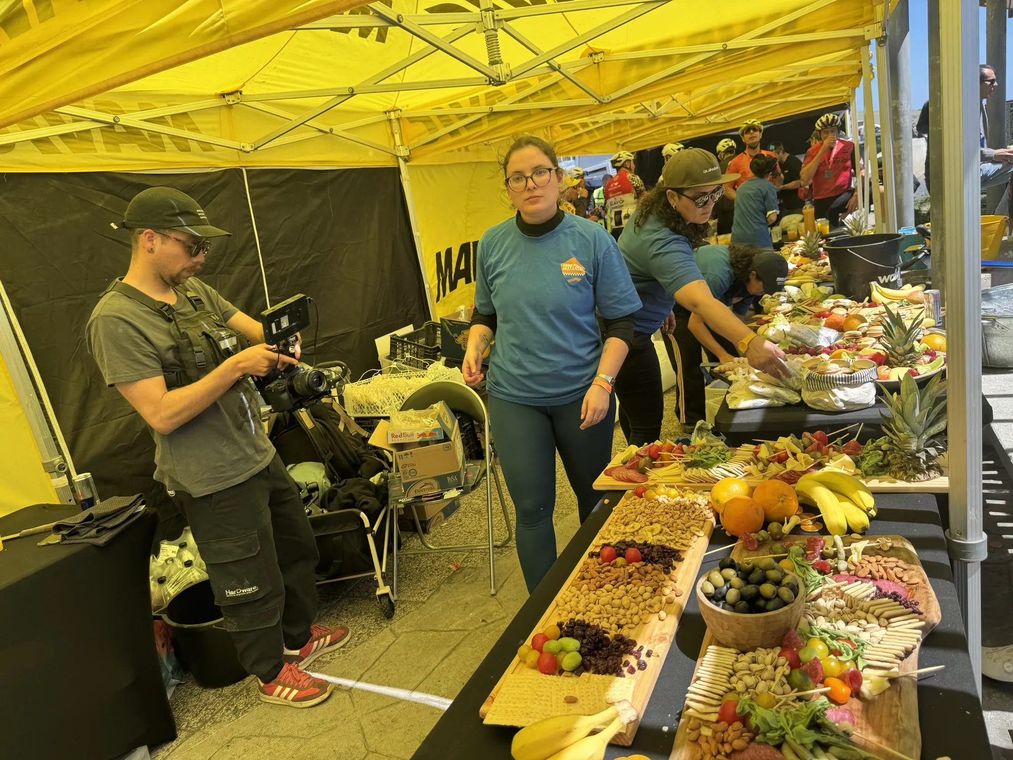 Mesas con picoteo para los corredores en el tercer tiempo: frutos secos, frutas, quesos y piñas bajo un toldo amarillo, con el staff atendiendo
