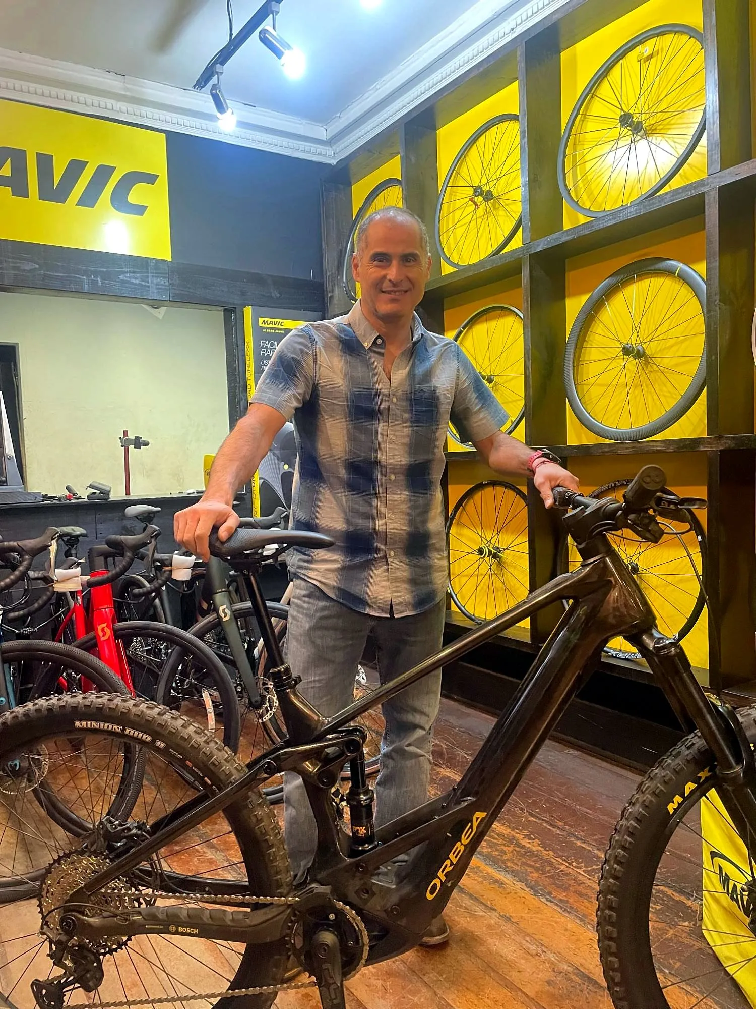Cliente junto a una eMTB en una tienda especializada, con un muro de ruedas y bicis de ruta al fondo