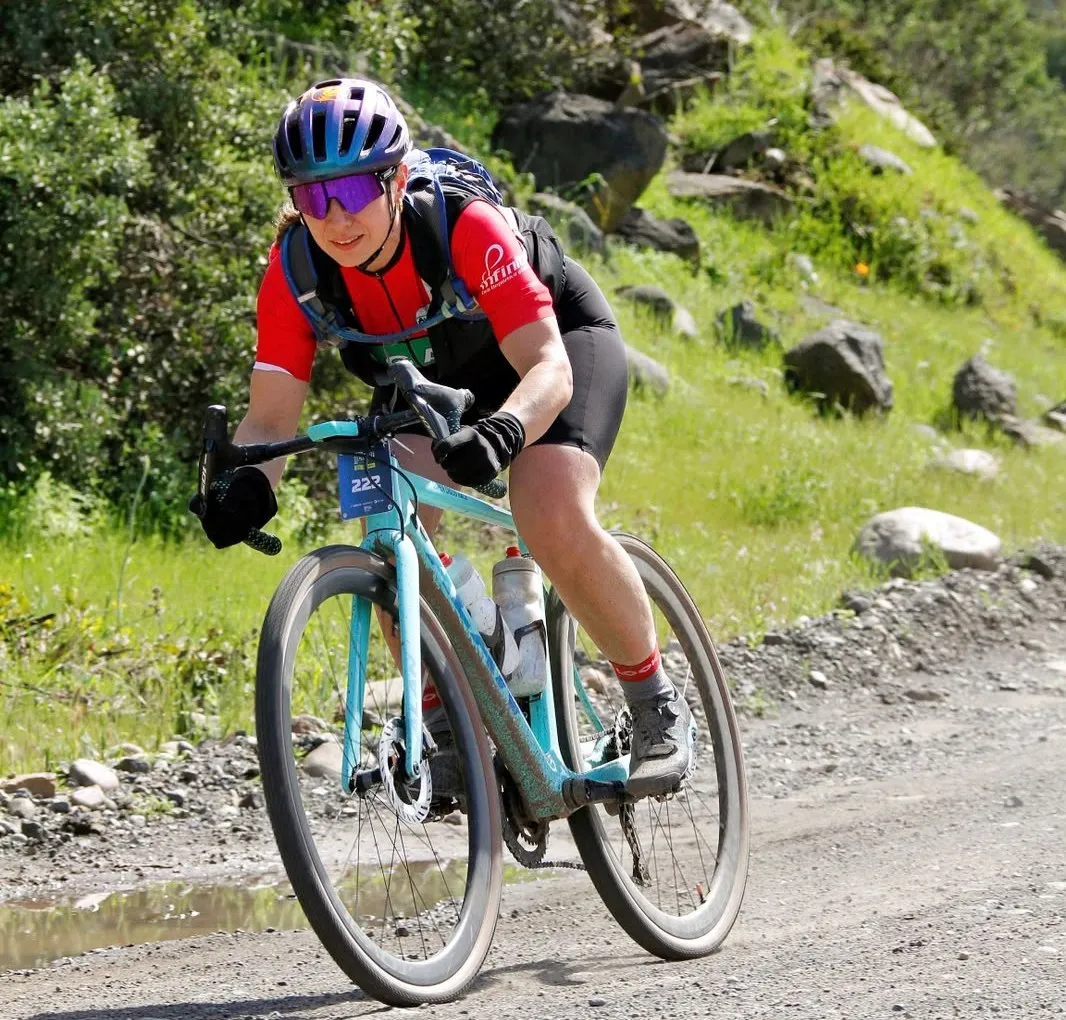 Verónica en su bicicleta de gravel, inclinada sobre el manubrio en un tramo de ripio con piedras y vegetación