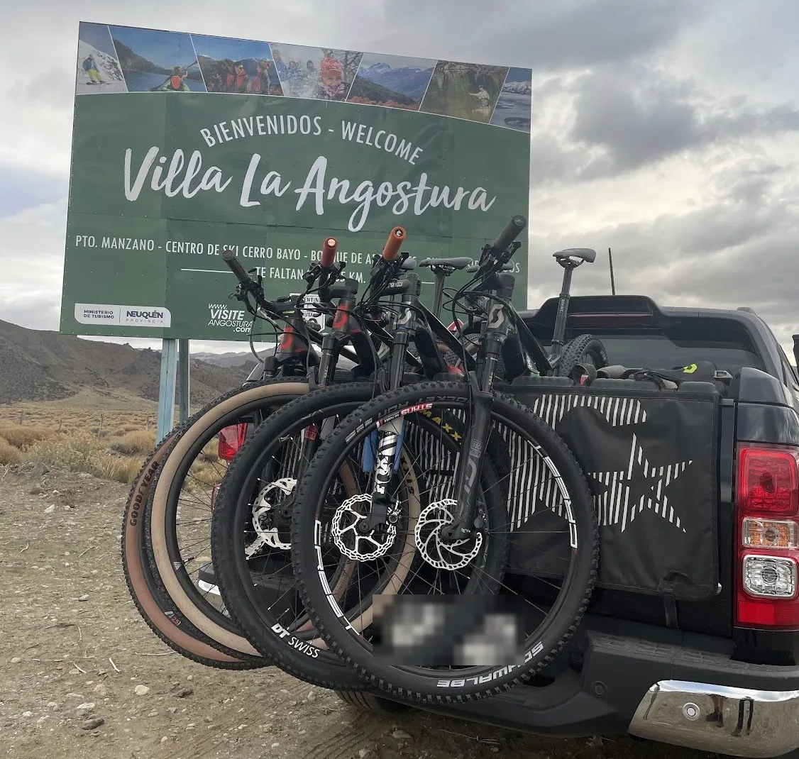 Cuatro bicis de montaña con suspensión colgadas del pad Evoc en la camioneta, frente al letrero de bienvenida a Villa La Angostura, en Argentina