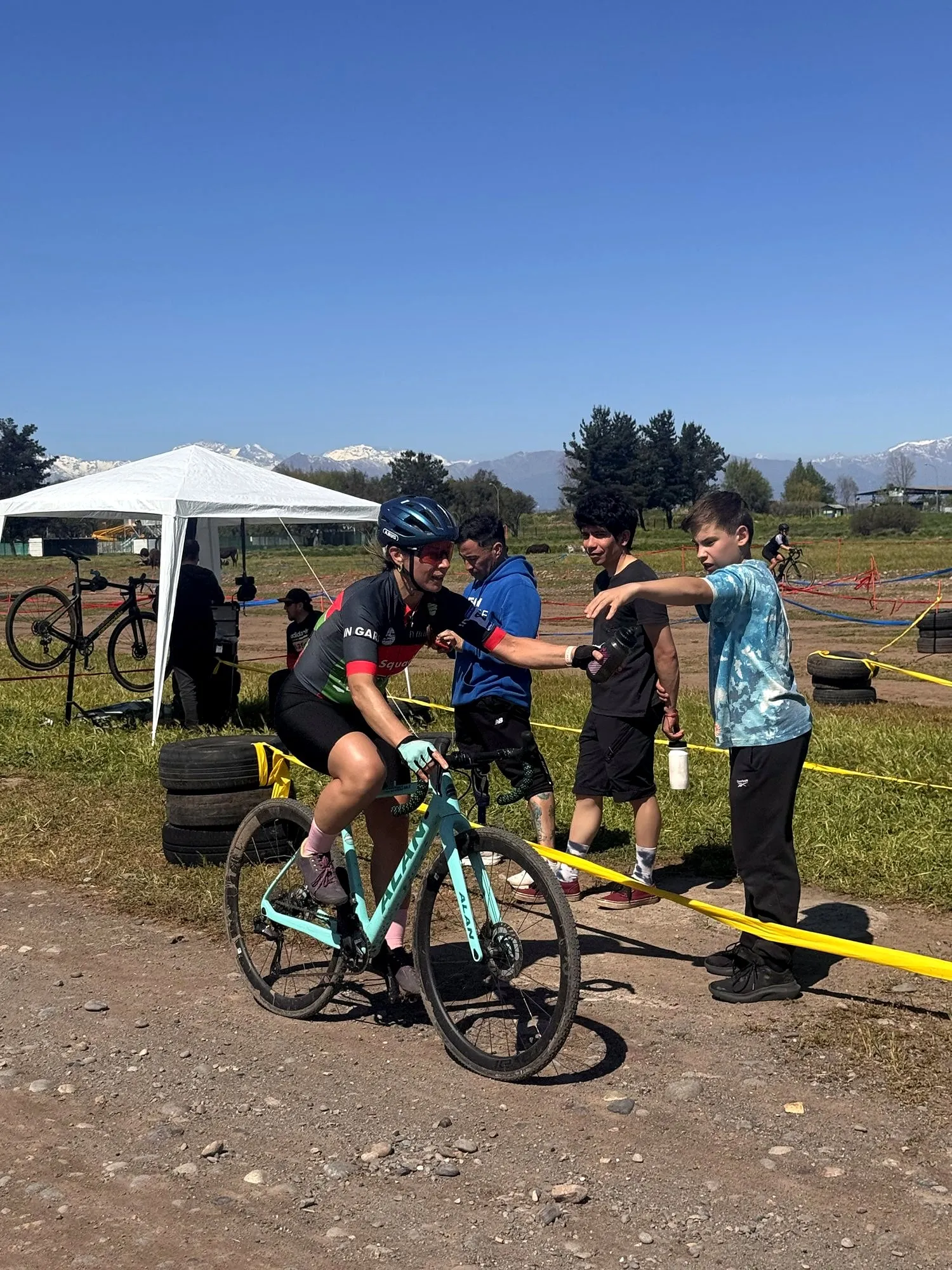 Ciclista de ciclocross recibiendo una caramayola desde la zona de abastecimiento durante una carrera, con la cordillera de fondo